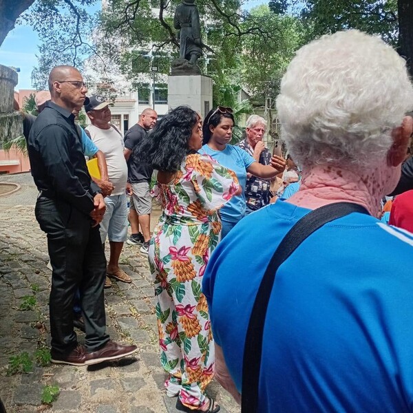 Grupo de pessoas em um parque ou praça, algumas ouvindo atentamente uma mulher de vestido floral que parece estar falando ou explicando algo.