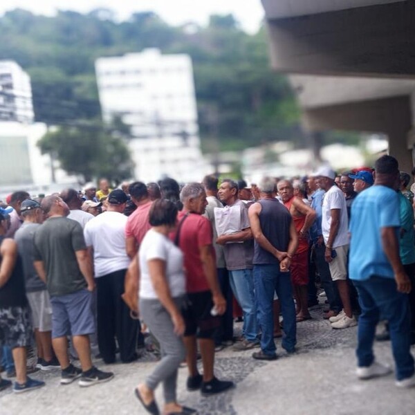 Multidão de pessoas reunidas em uma calçada, algumas conversando entre si, com prédios e vegetação ao fundo.