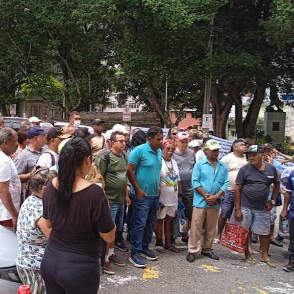 Grupo de pessoas reunidas ao ar livre, algumas de pé e outras conversando, em um ambiente com árvores e edifícios ao fundo.
